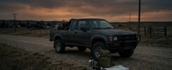 Pickup truck on a rural gravel road at dusk, with a backpack, water container, flashlight, and map in the foreground.