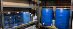 Blue water-storage barrels, dated containers and bottled water arranged on basement shelving.